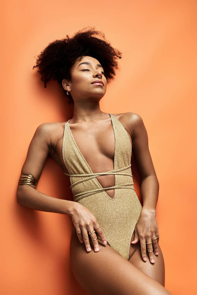Stylish African American woman in gold swimsuit strikes a pose on vibrant orange backdrop. - Photo, Image