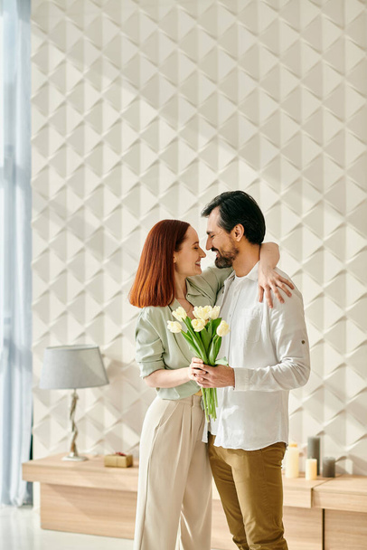 Una mujer pelirroja y un hombre barbudo se paran frente a una pared con flores en un apartamento moderno, disfrutando de un tiempo de calidad juntos. - Foto, Imagen