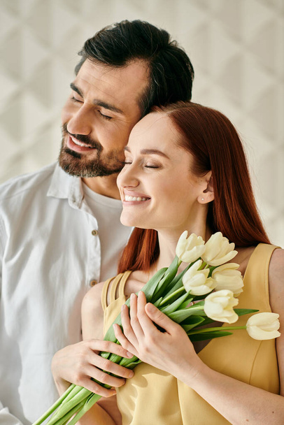 Una rossa e un uomo barbuto, con in mano un mazzo di tulipani, che condividono un momento di tenerezza in un appartamento moderno. - Foto, immagini