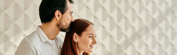 Egy gyönyörű felnőtt pár, egy vörös hajú nő és egy szakállas férfi, akik egy fehér fal előtt ölelkeznek a modern lakásukban.. - Fotó, kép