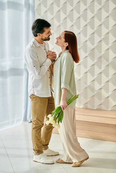 Kızıl saçlı bir kadın ve sakallı bir adam rengarenk çiçeklerle süslenmiş bir odada sarılıp evde birlikte güzel vakit geçiriyorlar.. - Fotoğraf, Görsel