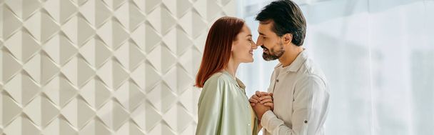 Пара, рыжая женщина и бородатый мужчина, целуются перед белой стеной в современной квартире. - Фото, изображение
