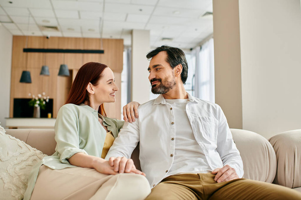 Рыжая женщина и бородатый мужчина сидят на стильном диване, наслаждаясь компанией друг друга в современной гостиной. - Фото, изображение
