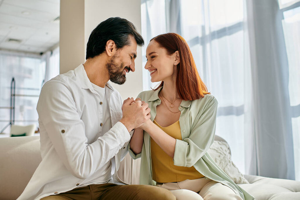 Ein schönes erwachsenes Paar, eine rothaarige Frau und ein bärtiger Mann sitzen zusammen auf einer Couch in einem modernen Appartement und genießen die schöne Zeit. - Foto, Bild