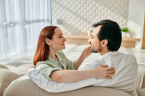 Egy gyönyörű felnőtt pár, egy vörös hajú nő és egy szakállas férfi ül szorosan a kanapén a modern nappaliban.. - Fotó, kép