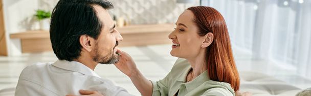 Una mujer pelirroja y un hombre barbudo comparten un momento tierno mientras se miran a los ojos en un apartamento moderno. - Foto, imagen
