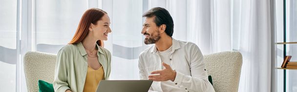 Een man met baard en een roodharige vrouw die een diep gesprek voeren terwijl ze comfortabel op een moderne bank zitten in een gezellig appartement. - Foto, afbeelding