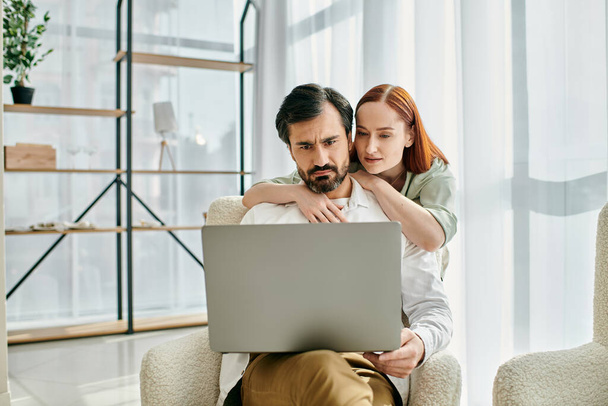 Een roodharige vrouw en een bebaarde man zitten samen in een stoel, werken aan een laptop in een modern appartement. - Foto, afbeelding