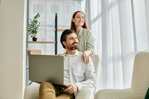 Een roodharige vrouw en een bebaarde man zitten nauw in een stoel, verdiept in het gebruik van een laptop samen in hun moderne appartement. - Foto, afbeelding