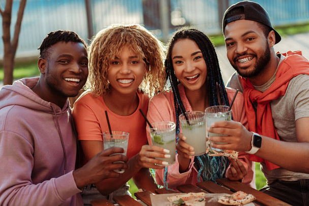 Grupo de diversos amigos afroamericanos sonrientes, reunidos en la fiesta de cumpleaños, sentados en la cafetería, bebiendo limonada y mirando a la cámara al aire libre. Concepto de celebración  - Foto, imagen