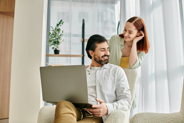 Pelirroja mujer y barbudo hombre sentado juntos, centrado en la pantalla del ordenador portátil en apartamento moderno. - Foto, Imagen