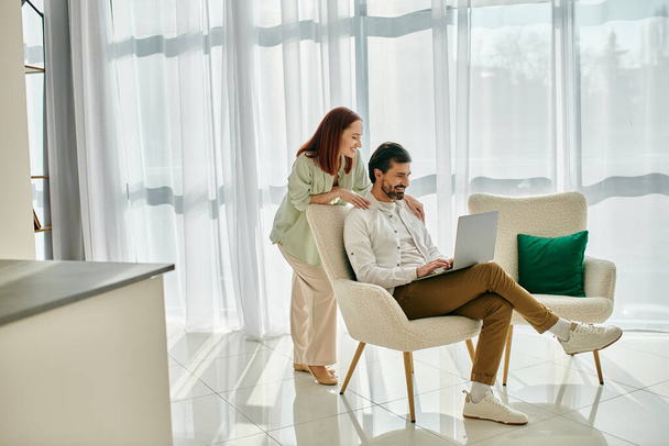 Een roodharige vrouw en een bebaarde man zitten in een stoel, met behulp van een laptop samen in een modern appartement, genieten van quality time. - Foto, afbeelding