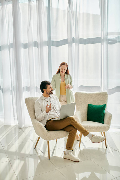 Un homme barbu et une femme rousse, assise sur une chaise avec ordinateur portable, profitent d'un moment de qualité dans un appartement moderne. - Photo, image