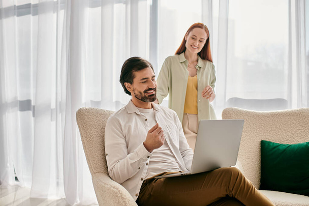 Een roodharige vrouw en een man met baard zitten samen op een bank, met een laptop in een modern appartement.. - Foto, afbeelding