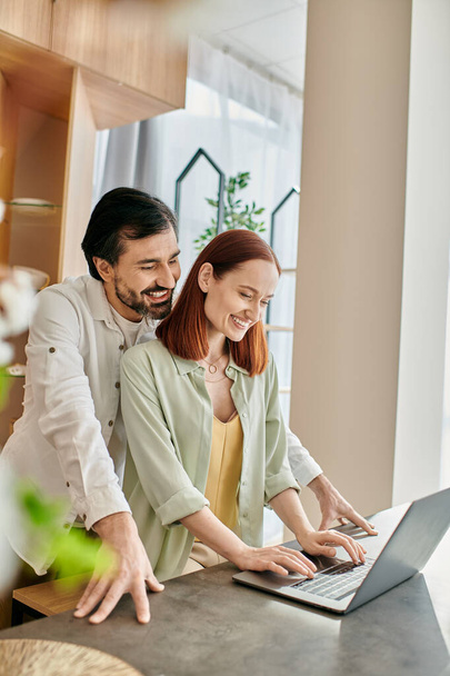 A redhead woman and bearded man engrossed in the screen of a laptop while standing in their modern kitchen. - Photo, Image