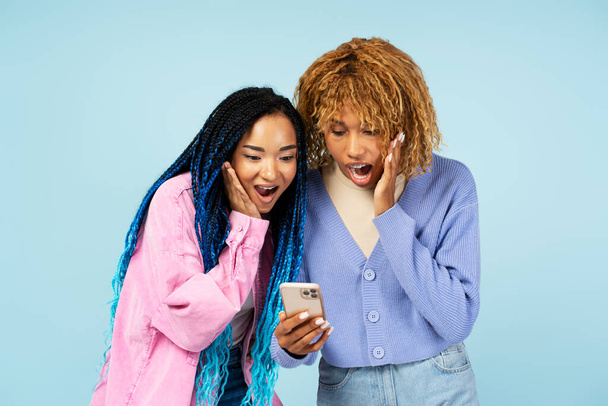 Muotokuva ikionnellinen naisten ystäviä tilalla matkapuhelin ostoksia verkossa myynti, vedonlyönti, juhla menestys eristetty sinisellä taustalla - Valokuva, kuva