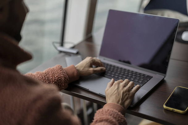 Over-de-schouder zicht van de handen van een man typen op een laptop naast een smartphone op een bureau met een uitzicht op zee. - Foto, afbeelding