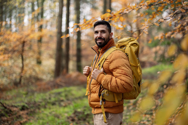 Веселий бородатий пішохід у жовтій куртці насолоджується подорожжю серед осіннього листя. - Фото, зображення