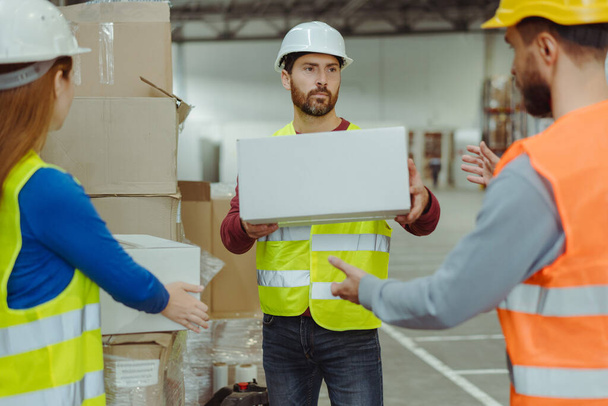 Handsome middle aged worker wearing hard hat and uniform, holding box, talking with colleagues in warehouse. Concept of teamwork, cooperation - Photo, Image