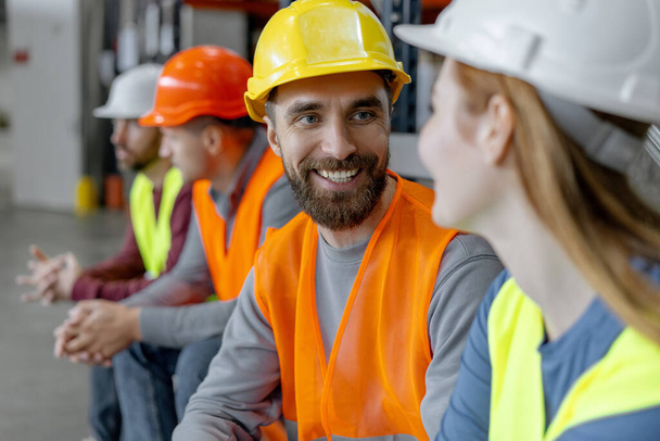 Positive colleagues wearing protective helmets and work wear, work in warehouse, talking during break. Smiling bearded worker, manager. Concept of teamwork, communication - Photo, Image