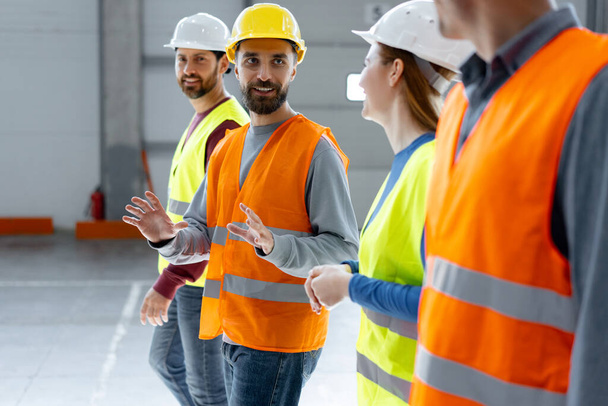 Group of confident, successful, happy managers wearing hard hats and vests, walking through large warehouse, discussing cooperation, distribution, planning. Teamwork concept - Photo, Image