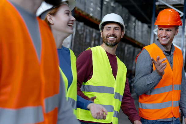 Group of smiling managers wearing hard hats and vests, walking through large warehouse, discussing cooperation, distribution, planning. Teamwork concept - Photo, Image