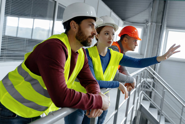 Successful, factory workers, managers wearing hard hats, vests and work wear talking, planning work, looking away in warehouse. Concept of industry, logistics, business - Photo, Image