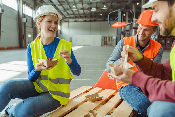 Trabajadores felices y sonrientes, gerentes de fábrica con sombreros duros, chalecos comiendo comida, sentados, hablando mientras almorzaban juntos en el almacén. Concepto alimenticio - Foto, Imagen