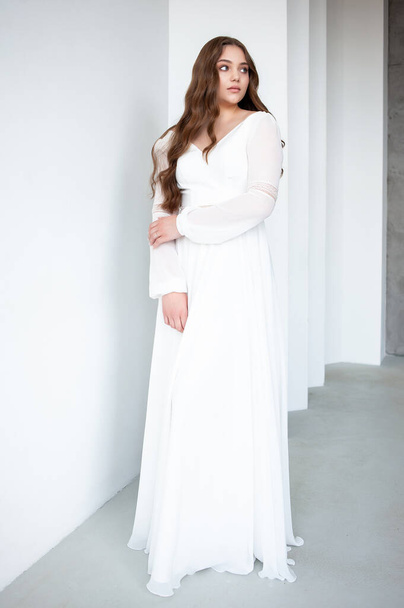 mooi meisje met lang haar in witte trouwjurk met een wit kapsel en een sluier op een licht grijze achtergrond - Foto, afbeelding