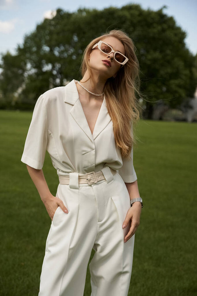 A stylish woman in a white jumpsuit strikes a pose in a vast, green field surrounded by serenity and beauty. - Photo, Image