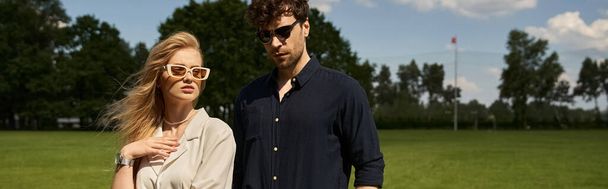 Un uomo e una donna in occhiali da sole, che indossano abiti eleganti, si trovano in un lussureggiante campo verde, trasudando stile old-money e uno stile di vita lussuoso. - Foto, immagini