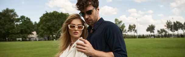 Stylový pár ve slunečních brýlích stojí na svěžím poli, vyzařuje eleganci a důvěru v jejich módní oblečení. - Fotografie, Obrázek
