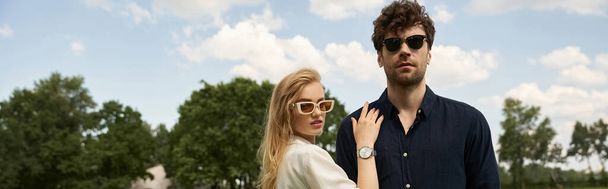 Un uomo e una donna alla moda, vestiti elegantemente, stanno insieme in un vivace campo verde, entrambi indossando occhiali da sole.. - Foto, immagini