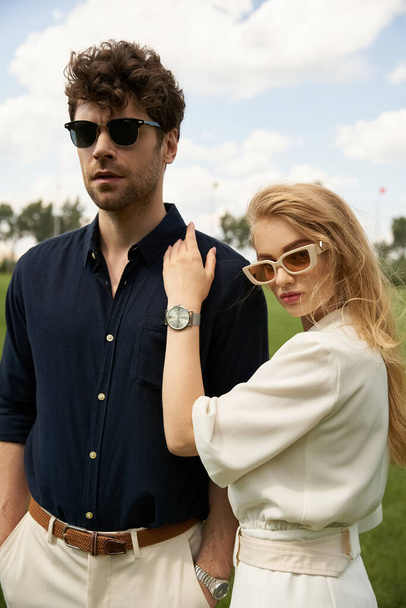 Un hombre y una mujer elegantes y ricos que lucen gafas de sol en un campo exuberante, exudando un aire de sofisticación y lujo. - Foto, imagen