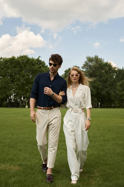 Een man en vrouw in elegante kleding genieten van een ontspannen wandeling in een weelderig, groen veld. - Foto, afbeelding