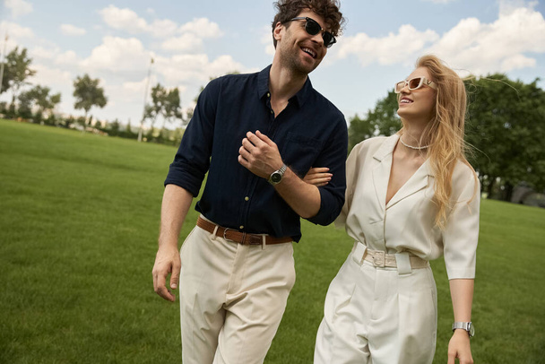 A beautiful young couple dressed in elegant clothing enjoys a leisurely walk in the park on a green field, embodying old money style and rich people lifestyle. - Photo, Image