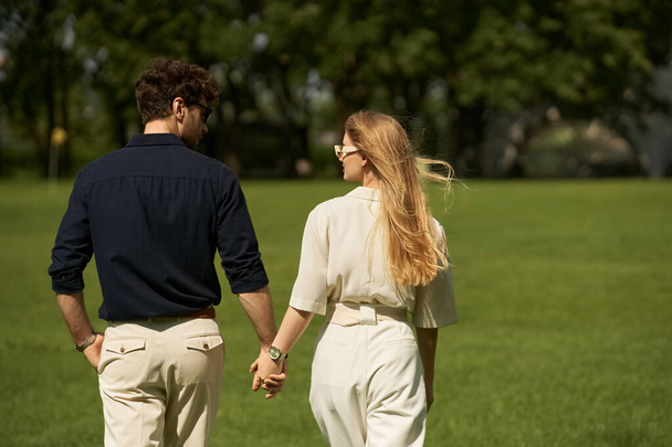 Een mooi jong echtpaar in elegante kledij hand in hand tijdens een wandeling door een park op een zonnige dag. - Foto, afbeelding