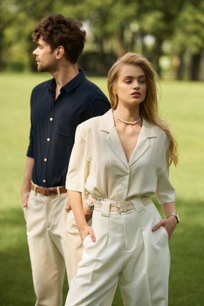Krásný mladý pár v elegantním oblečení stojí bok po boku v rozlehlém travnatém poli, vyzařuje nadčasovou sofistikovanost. - Fotografie, Obrázek
