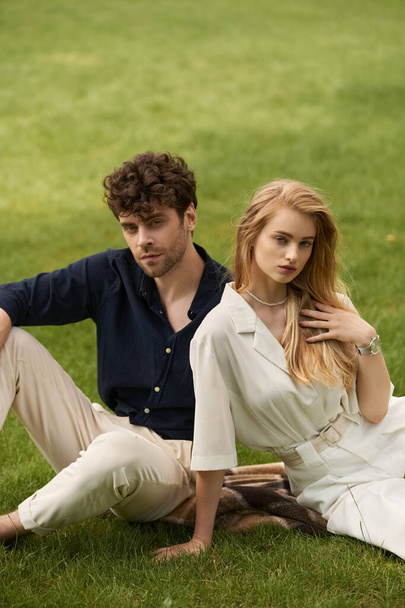 Ein stilvolles Paar in eleganter Kleidung sitzt zusammen auf grünem Gras und genießt die Gesellschaft des anderen in einer ruhigen Outdoor-Umgebung. - Foto, Bild
