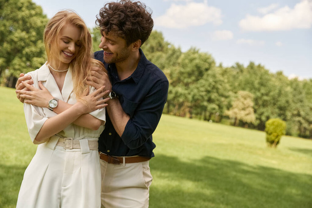 Красивая молодая пара в элегантной одежде нежно обнимается в зеленом поле, воплощая любовь и спокойствие. - Фото, изображение