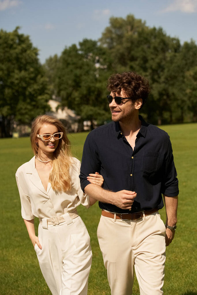 Una pareja joven y elegante vestida con ropa fina paseando tranquilamente por un exuberante parque verde en un día soleado. - Foto, imagen