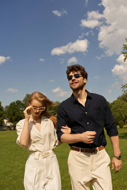 Un beau jeune couple en vêtements élégants se promène dans un parc verdoyant, incarnant un style old-money et un style de vie luxueux. - Photo, image
