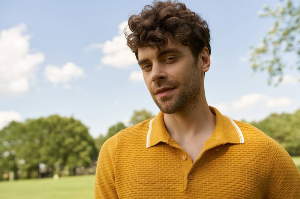 Een stijlvolle man in een levendig geel shirt staat midden in het weelderige groen van een park op een zonnige dag. - Foto, afbeelding
