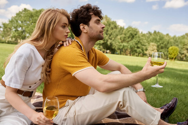 Couple élégant profitant d'un pique-nique romantique dans un parc, tenant des verres à vin et immergé dans la conversation. - Photo, image