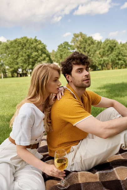 Ένα κομψό νεαρό ζευγάρι, κομψά ντυμένο, χαλαρώνει σε μια κουβέρτα σε ένα καταπράσινο πάρκο, ενσωματώνοντας έναν πολυτελές τρόπο ζωής. - Φωτογραφία, εικόνα