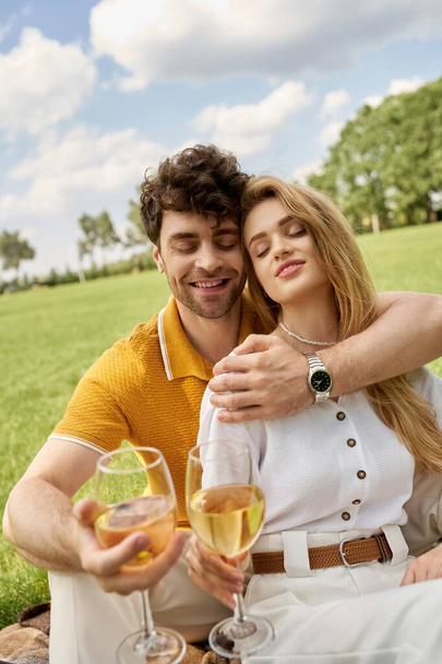 Красивая молодая пара в элегантной одежде тосты с бокалами вина в пышном парке, воплощая роскошь и изысканность. - Фото, изображение
