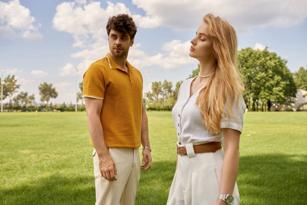Красивая молодая пара в элегантной одежде, стоящая вместе в зеленом поле, олицетворяя старинный стиль денег и роскошный образ жизни. - Фото, изображение
