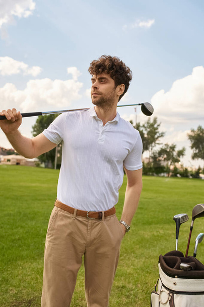 Egy elegáns ruhás férfi golftáskát és ütőt tart egy buja zöld mezőn egy tekintélyes golfklubban.. - Fotó, kép