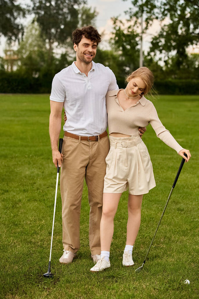 Un hombre joven y una mujer en traje elegante posan amorosamente en un campo de golf verde bajo el cielo despejado. - Foto, imagen