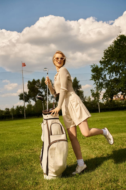 Μια γυναίκα με κομψή ενδυμασία στέκεται σε ένα χλοερό πεδίο με μια τσάντα του γκολφ, έτοιμη να παίξει μια ηλιόλουστη μέρα στο γκολφ κλαμπ. - Φωτογραφία, εικόνα